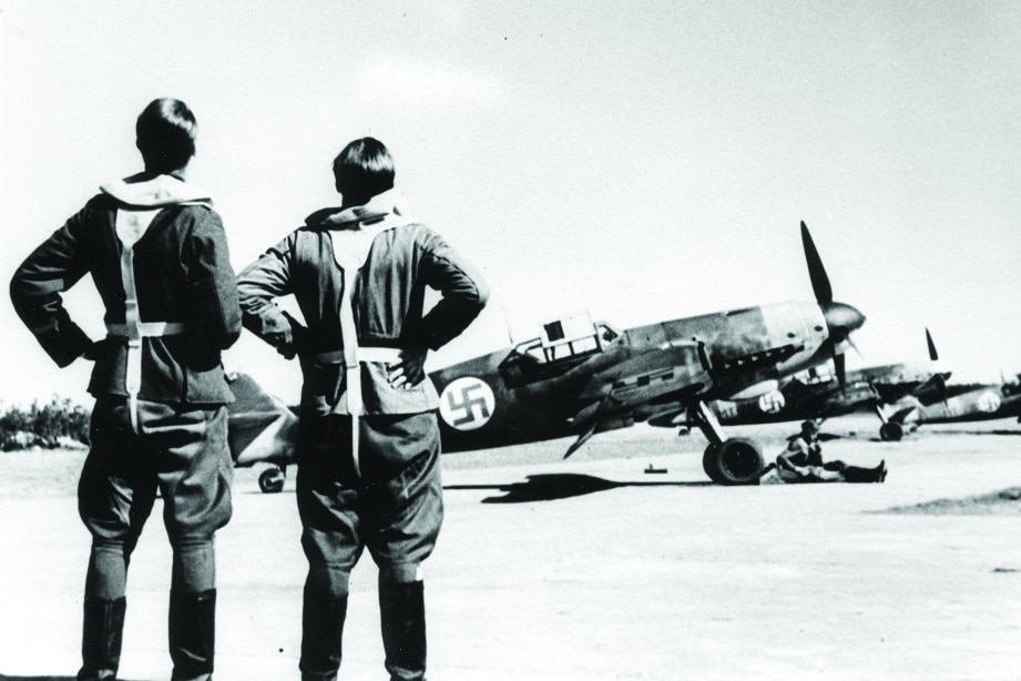 Vanha kuva, jossa kaksi lentäjää seisoo lentokentän reunalla katsellen Messerschmitt-hävittäjiä.